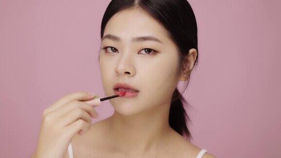 特写脸美丽的年轻亚洲女人应用嘴唇在她的嘴唇粉红色的背景