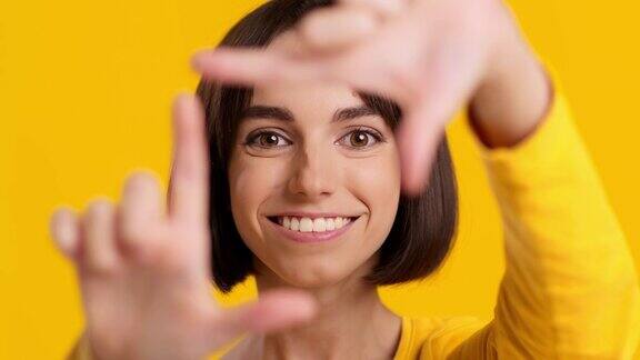黄色背景年轻女子透过手指相框看相机