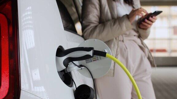 女人用手机附近充电的电动汽车;在户外公共充电站充电的车辆汽车共享