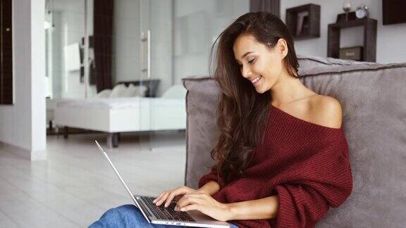 快乐的黑发女人在毛衣坐在地板上和使用笔记本电脑的侧面视图