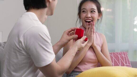 快乐的亚洲男人把装在小红盒子里的订婚戒指送给家里客厅里的女人