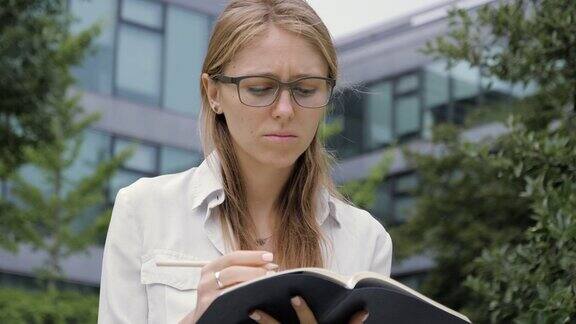 年轻的女学生戴着眼镜坐在户外的长椅上写笔记