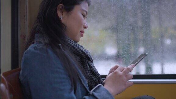 年轻快乐的亚洲女性在旅行期间坐在公共交通穿梭巴士的窗户边使用智能手机而外面下雨女性在欧洲度假旅行