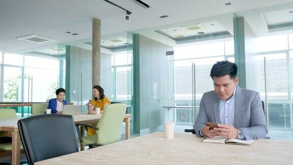 亚洲商人的肖像工作与智能手机坐在现代办公室商务人士的同事在背景