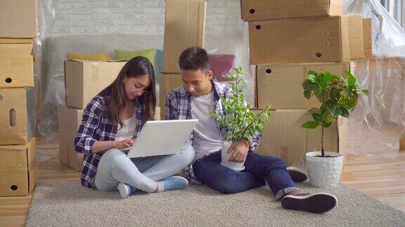 一对年轻的亚洲夫妇开车进入一个新公寓坐在地板上背景里放着盒子使用笔记本电脑