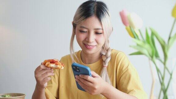 年轻的亚洲女子愉快地吃着披萨在家里用手机亚洲休闲用餐