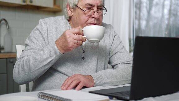 戴眼镜的老年男性企业主坐在笔记本电脑前的办公桌前在家工作的成功年长男性灰色的头发60-70岁的老忙人打字键盘看显示器距离办公室