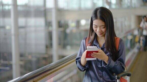 年轻的亚洲女子在机场候机楼的自动扶梯上检查手机女性在旅途中提着行李女孩用智能手机聊天微笑着等飞机旅行