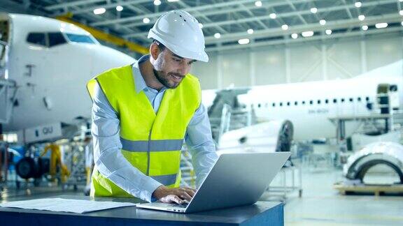 飞机开发工程师穿着安全背心和安全帽使用笔记本电脑和蓝图分析检查和工作飞机设计设施