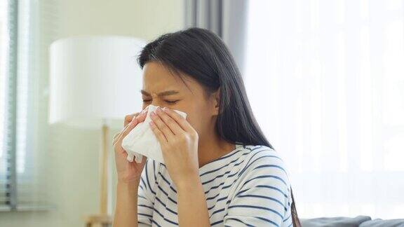 亚洲美女在客厅流鼻涕和打喷嚏有魅力的年轻女性坐在沙发上感觉不好患上了过敏然后在家里打喷嚏时用纸巾捂住鼻子