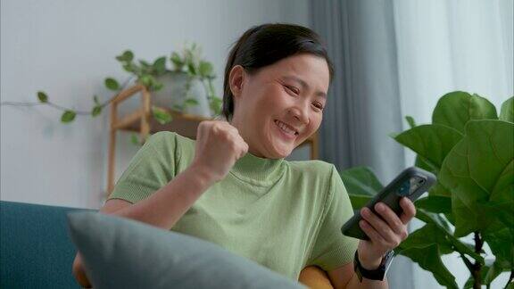 亚洲女人坐在家里客厅的沙发上看着智能手机开心兴奋地赢得了比赛