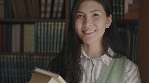 无忧无虑的亚洲女学生站在大学图书馆拿着书微笑着看着镜头