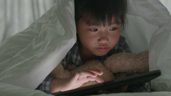 亚洲小女孩晚上在卧室里盖着毯子用平板电脑