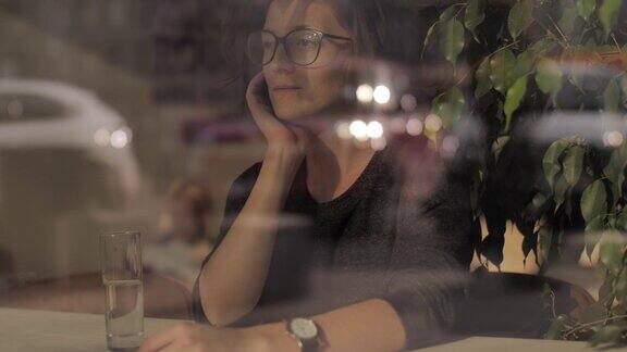近距离拍摄的一个年轻迷人的女孩在眼镜坐在咖啡店通过窗口视图路过的人都在反思
