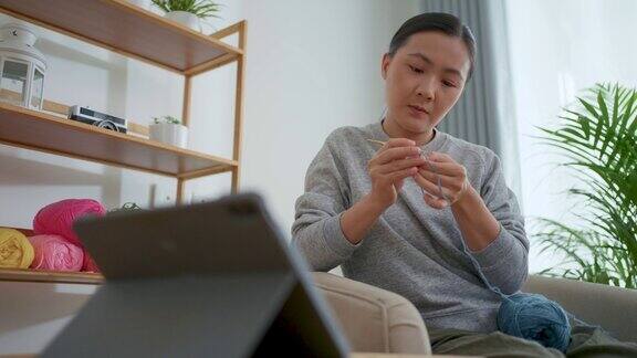 亚洲妇女坐在家里的客厅里从平板电脑上的在线视频剪辑中学习如何钩针编织