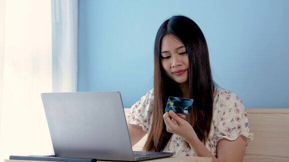 4K50帧特写美丽的亚洲女孩拿着蓝色信用卡填写信用卡信息坐在网上购物通过笔记本电脑使用信用卡在线支付