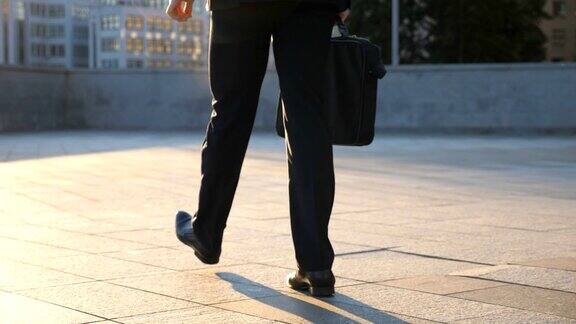 一个年轻商人的脚拿着公文包走在城市的街道上通勤上班的商人穿着西装的自信男人正在上班的路上城市景观背景慢镜头后视图特写