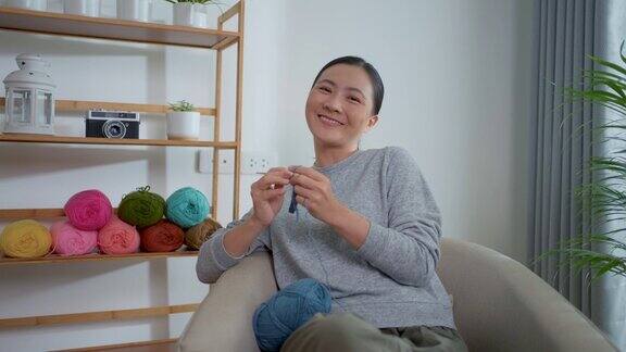 一个亚洲女人坐在家里客厅的扶手椅上看着相机一边编织钩针