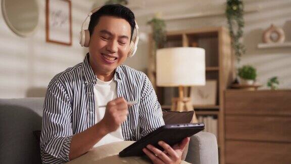 幸福亚洲成年男性自由职业者微笑愉快花时间工作平板电脑耳机社交媒体娱乐有趣的内容流休息在家里的沙发客厅周末度假