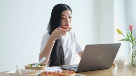 年轻的亚洲妇女吃披萨沙拉和意大利面同时在家里用笔记本电脑工作亚洲休闲餐饮