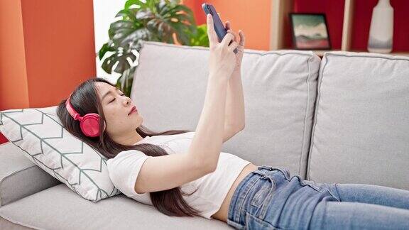 年轻的中国女子戴着耳机躺在家里的沙发上玩智能手机