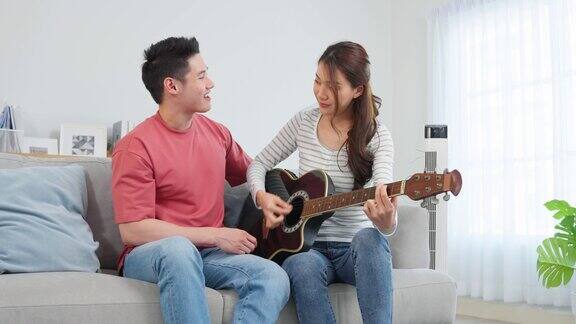 亚洲年轻情侣在家里的客厅里一起弹吉他迷人浪漫的新婚活跃浪漫的男人坐在沙发上教漂亮的女朋友在家里弹琴
