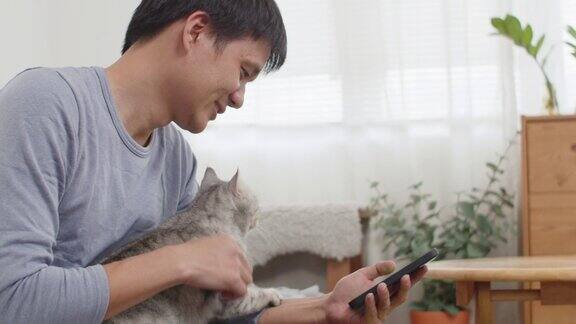 年轻的亚洲男子在家里用智能手机抚摸他可爱的猫