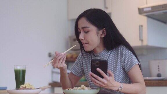 快乐的亚洲女人吃着亚洲面条泰式炒面在午休时间拿着筷子吃午饭在家厨房的桌子上打智能手机或浏览社交网络