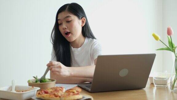 年轻的亚洲妇女吃披萨沙拉和意大利面同时在家里用笔记本电脑工作亚洲休闲餐饮