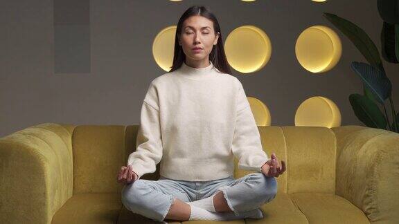 该上瑜伽课了有吸引力的年轻亚洲妇女训练和坐在一个瑜伽莲花姿势在沙发上