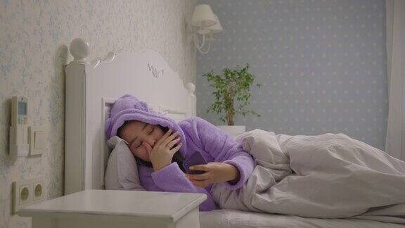 20多岁的亚洲女性穿着紫色睡衣躺在床上用手机昏昏欲睡的女人在床上用手机上网