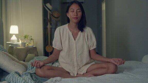 年轻的亚洲女人穿着睡衣她躺在床上咒语冥想晚上在家里的卧室里手拉手做瑜伽日常的生活方式