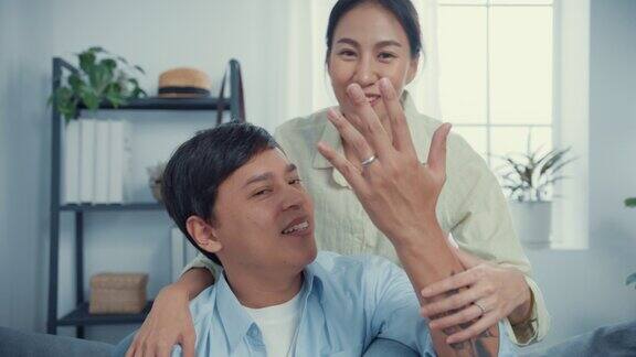年轻的亚洲夫妇与休闲坐在沙发上看相机显示美丽的戒指在客厅在家里