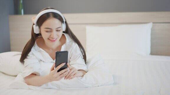 美丽的亚洲女人听耳机音乐与智能手机和聊天信息与她的男朋友微笑快乐在床上卧室舒适休息酒店人的生活方式