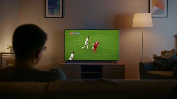 戴眼镜的年轻人正坐在沙发上看电视看足球赛现在是晚上家里的房间有工作灯