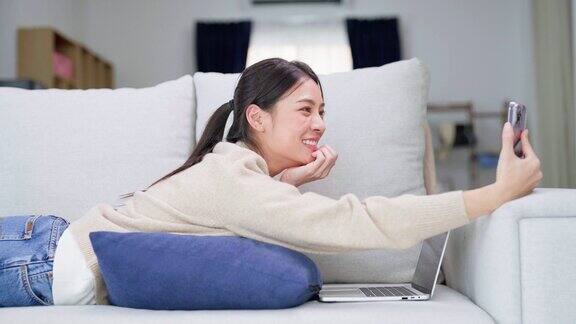 快乐的年轻亚洲女性在家里放松女人笑着躺在沙发上手里拿着手机女孩用视频打电话给朋友
