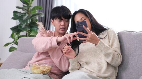 年轻的亚洲夫妇使用他们的手机和交谈同时坐在沙发上在家里客厅