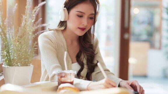 亚洲自由职业女性成人的微笑肖像一边用平板电脑远程在线工作一边听着耳机里的音乐心情愉悦积极向上在咖啡馆餐厅里自然光透过橱窗商店咖啡馆
