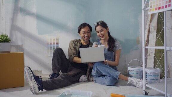 年轻的亚洲夫妇正在修补和粉刷墙壁他们坐在那里通过笔记本电脑查看家庭装修的想法