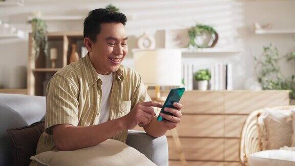 幸福亚洲成年男性丈夫微笑开朗花时间使用智能手机社交媒体娱乐有趣的内容流而休息在沙发上在家里的客厅周末假期