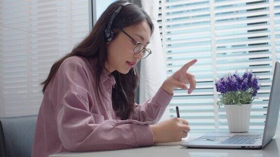 亚洲女学生在家桌上用笔记本电脑在线学习女性戴着耳机看着笔记本电脑说话解释和写笔记在家学习在线学习