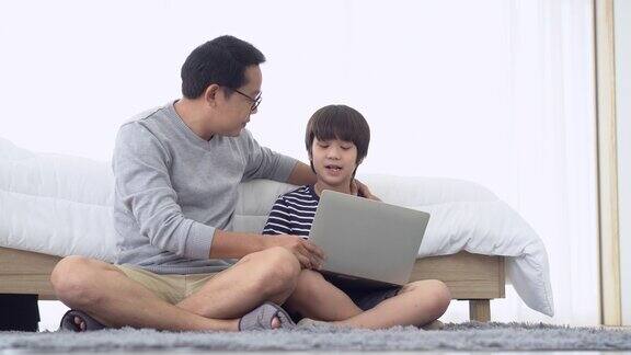 一位亚洲父亲坐在家里卧室的地板上一边用笔记本电脑做作业一边和可爱的儿子聊天在家学习和远程学习在线教育