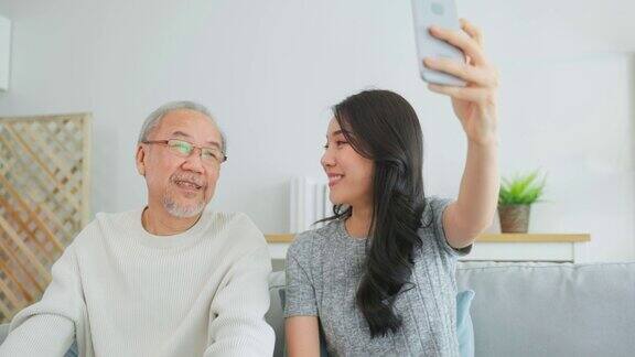 亚洲可爱的家庭女儿用电话与资深父亲视频通话年轻漂亮的女孩和年长的大龄男性坐在沙发上享受通过互联网交流在家里的客厅一起聊天