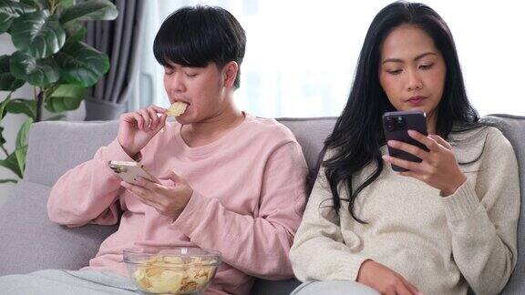 年轻的亚洲夫妇坐在沙发上玩他们的手机他们在专注于在线内容的同时忽略了彼此