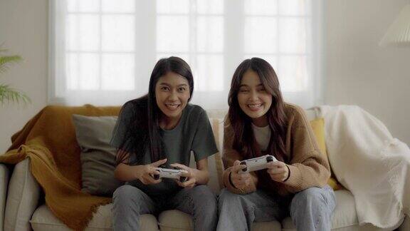 两个人漂亮年轻的亚洲女人坐在沙发上拿着操纵杆遥控器玩在线视频游戏在客厅