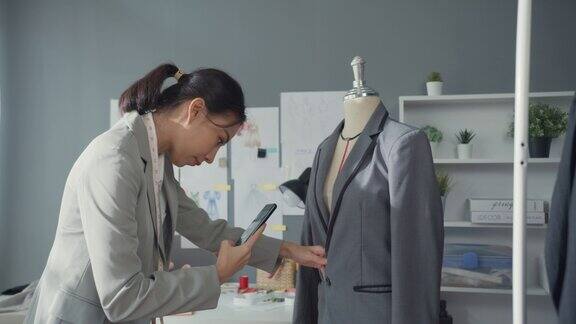 愉悦的亚洲女商人时装设计师带着套装系列坐在桌子上用智能手机拍照在工作室工作场所向客户确认采购订单启动业务