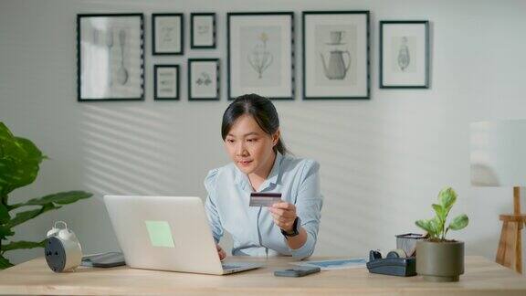 亚洲女性手持信用卡在家办公室用笔记本电脑进行网上支付