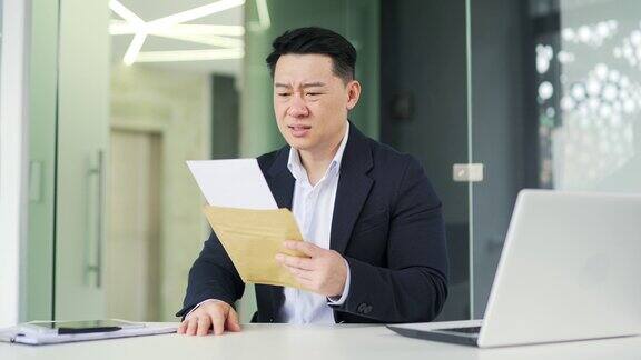 失望的亚洲人坐在办公室的办公桌前读着带坏消息的信