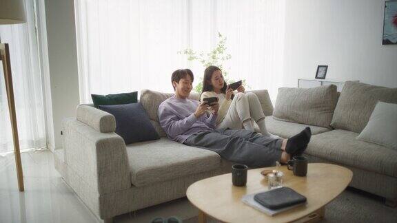 一对韩国夫妇坐在舒适的沙发上在时尚明亮的客厅里一起玩有趣的电子游戏一对美丽的情侣坐在家里的沙发上享受时光