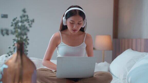 休闲放松亚洲年轻成年女性听音乐从耳机同时使用笔记本电脑技术设备在卧室工作在家亚洲女性工作在早上的日光在卧室周末例行时刻